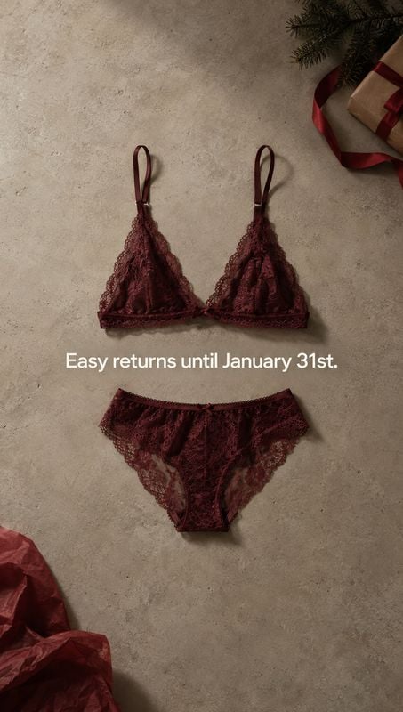 burgundy lace lingerie set flat lay product photo: A burgundy lace lingerie set is arranged on a textured neutral surface with festive greenery and gift wrap a…