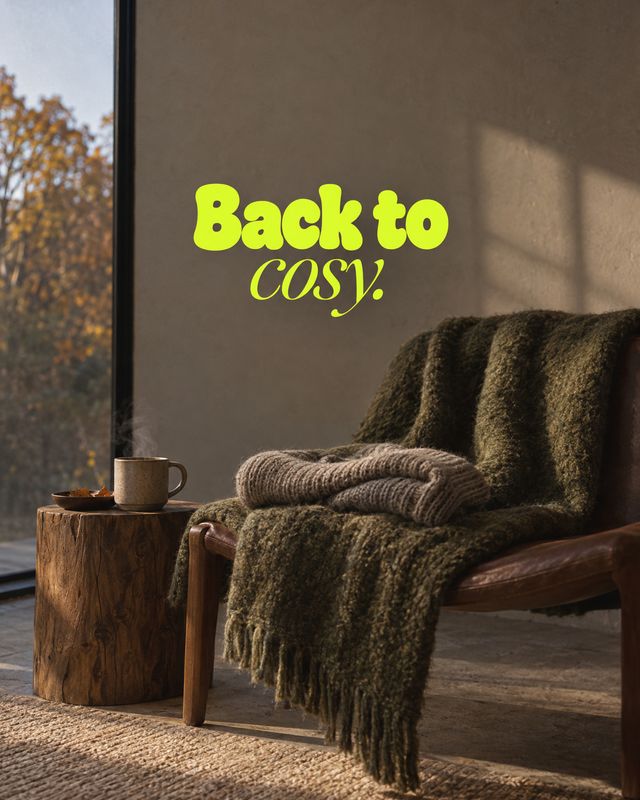 knitted throw blanket room set product photo: A chunky knitted throw drapes over a leather chair beside a wooden table and mug, with warm autumn light and brig…