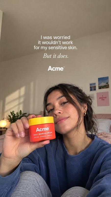 daily barrier cream product in hand product photo: A woman in a blue sweatshirt holds a bright red-and-yellow cream jar toward the camera in a softly sunlit be…
