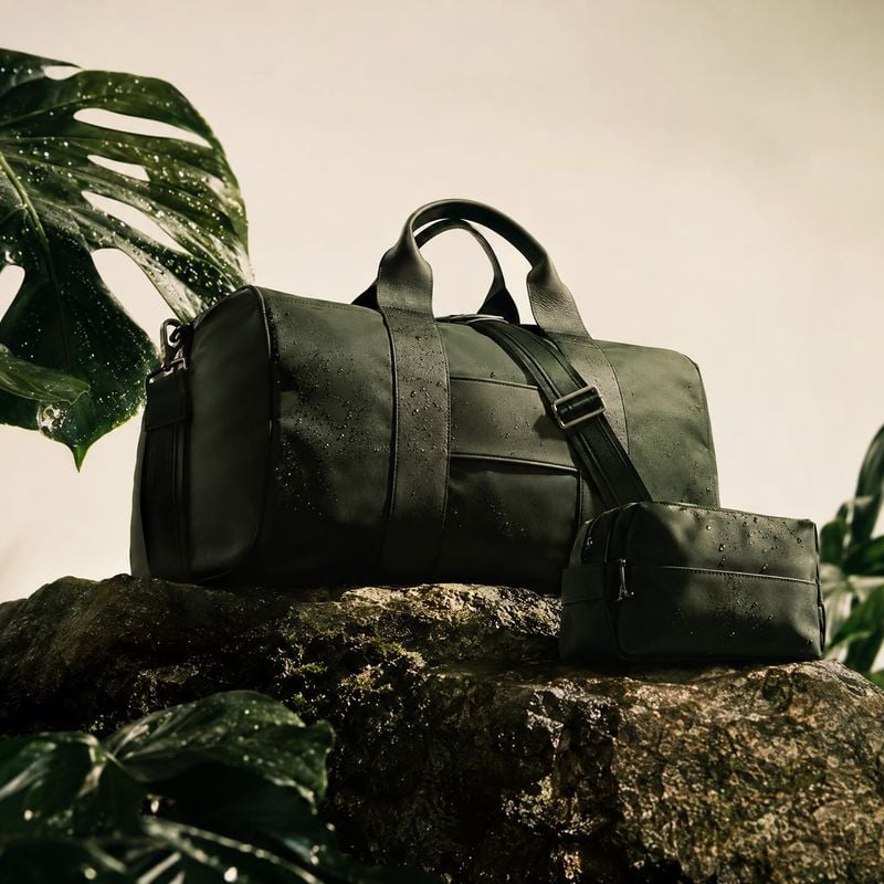 duffel bag and pouch lifestyle scene product photo: A dark green duffel bag and matching pouch rest on a mossy rock among tropical leaves, styled in warm natur…