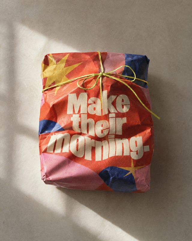 wrapped gift package flat lay product photo: A crinkled red, pink, and blue wrapped package tied with a thin gold ribbon sits against a warm beige background,…