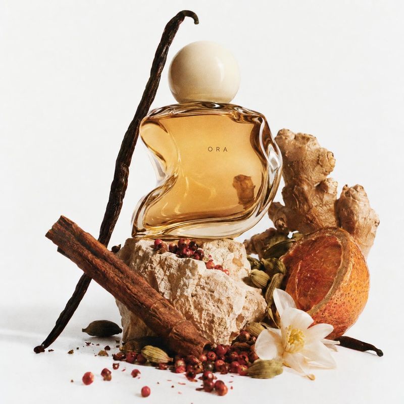 perfume ingredients product photo: An amber perfume bottle with a sculptural glass silhouette sits among vanilla, cinnamon, ginger, citrus and spices on a pale…