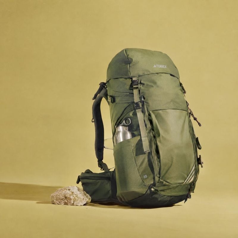 hiking backpack sports product photo: An olive-green hiking backpack with a water bottle and small rock is staged against a warm yellow seamless backdrop, with…