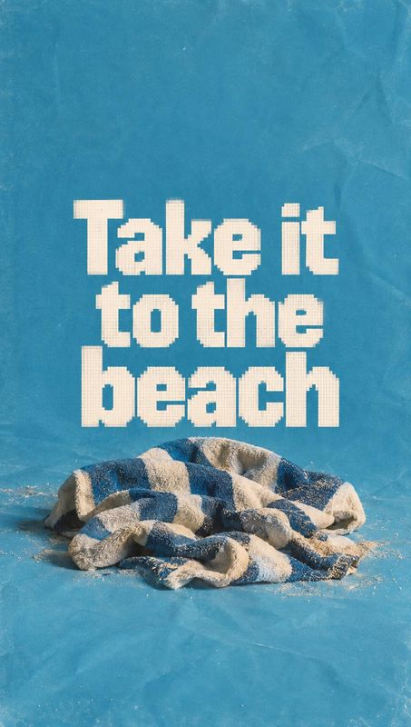 beach towel home product photo: A rumpled blue-and-beige beach towel rests on a sandy blue studio surface beneath oversized cream lettering, creating a playful…
