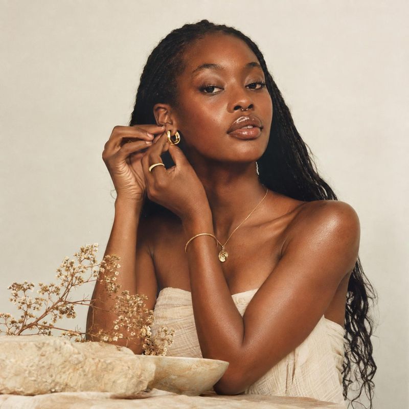 gold jewelry on-body detail product photo: A woman adjusts a gold hoop earring while wearing layered gold jewelry and a softly draped cream top against a warm…