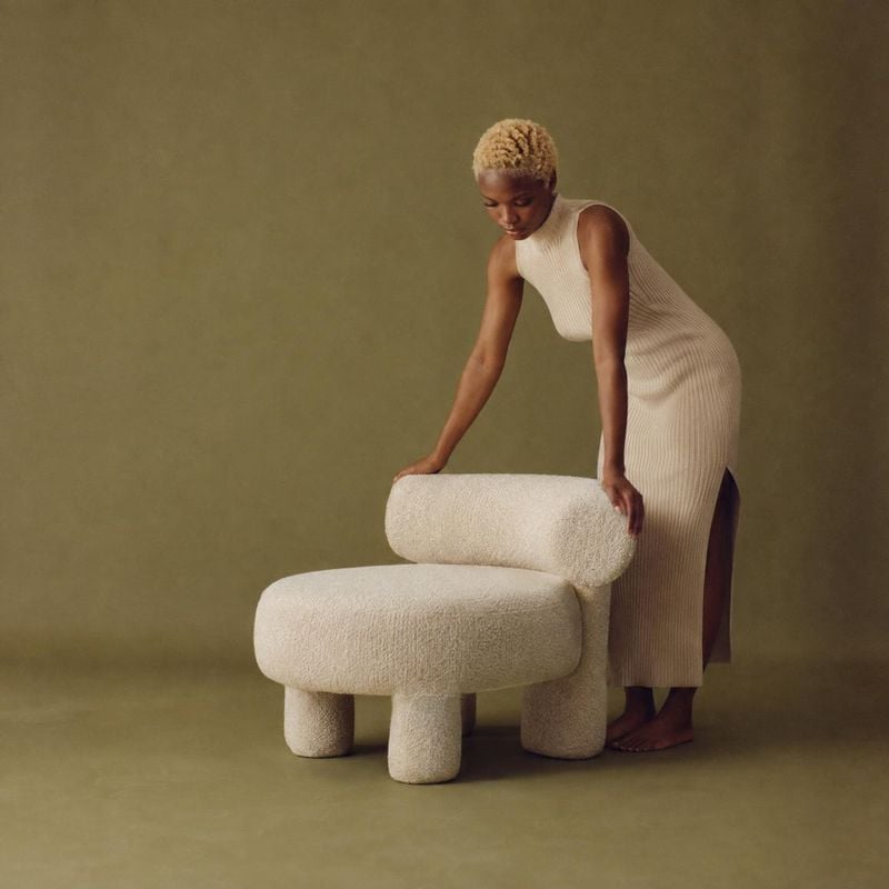upholstered accent chair lifestyle scene product photo: A woman in a cream ribbed dress leans over a sculptural ivory bouclé chair against a muted olive studio…