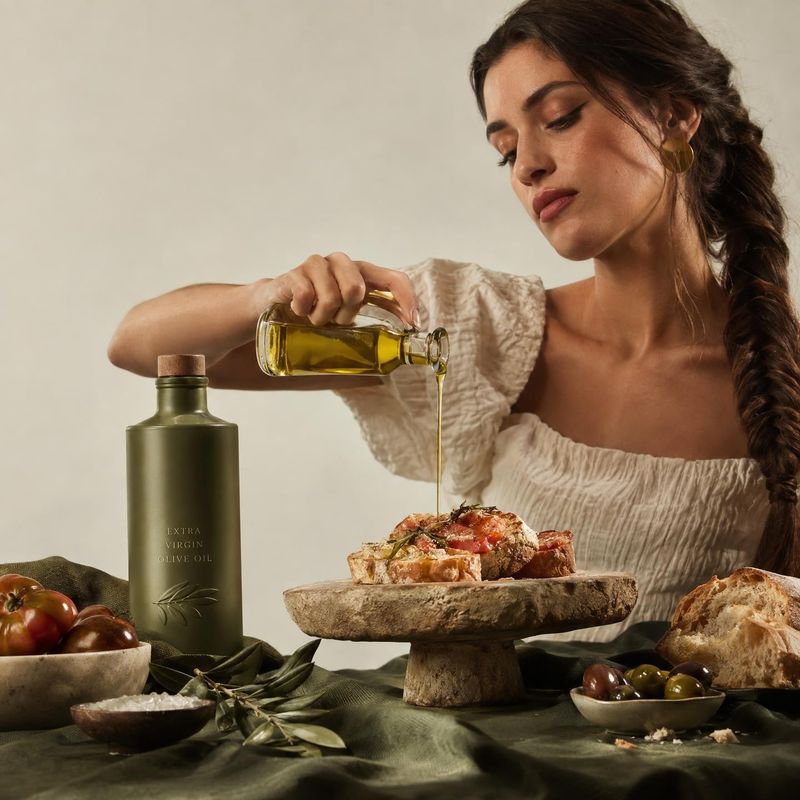 olive oil ingredients product photo: A woman in a cream blouse pours golden olive oil over rustic toast at a richly styled table, with warm directional light a…