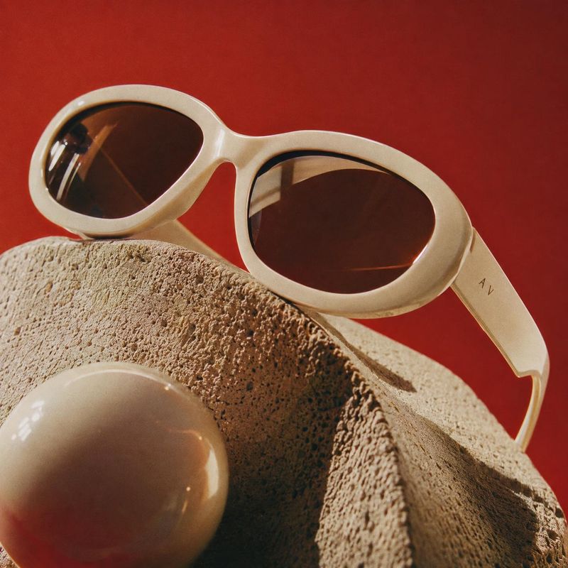 sunglasses close-up detail product photo: Cream-colored sunglasses rest diagonally across a rough sculptural stone form against a vivid red backdrop, with warm…