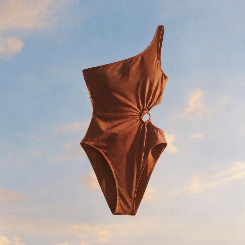 one-piece swimsuit floating product product photo: A rust-brown one-piece swimsuit with an asymmetrical strap and gathered ring detail floats against a softly…