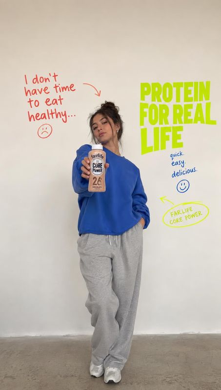 protein shake product in hand product photo: A model in a blue sweatshirt and gray sweatpants presents a protein shake against a plain studio wall, surrounded…