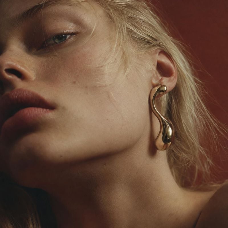 gold sculptural earring on-body detail product photo: A close, moody beauty portrait frames a model wearing a polished gold sculptural earring against a deep r…