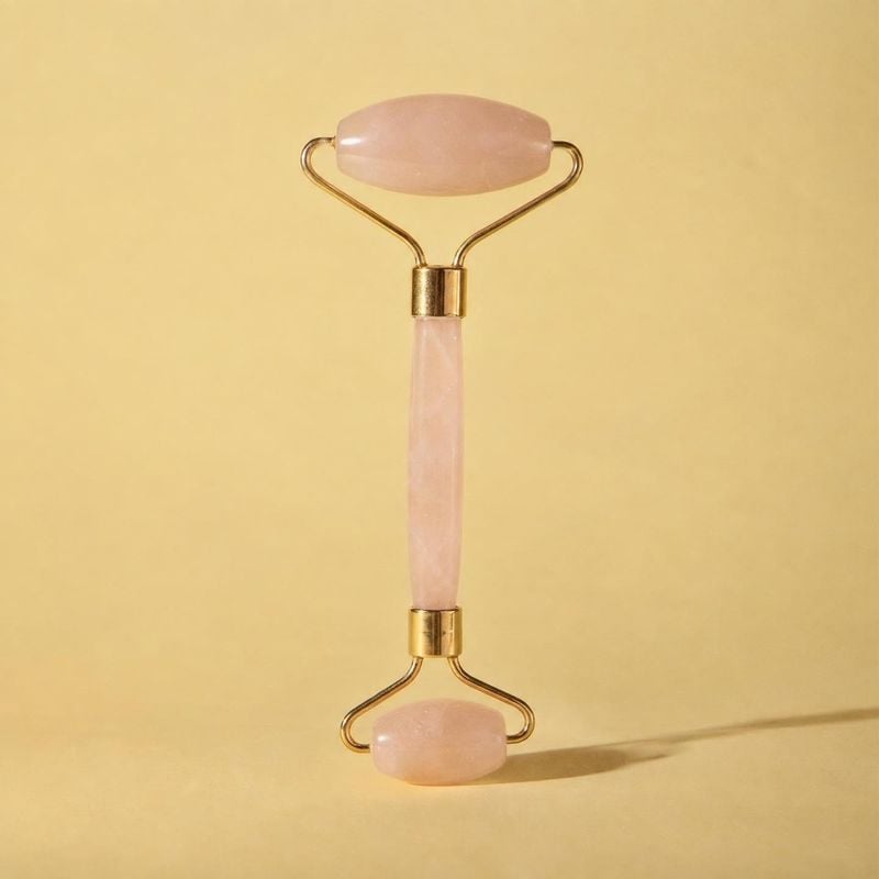 rose quartz facial roller beauty product photo: A rose quartz facial roller with gold-tone fittings stands upright against a warm yellow seamless backdrop, lit…