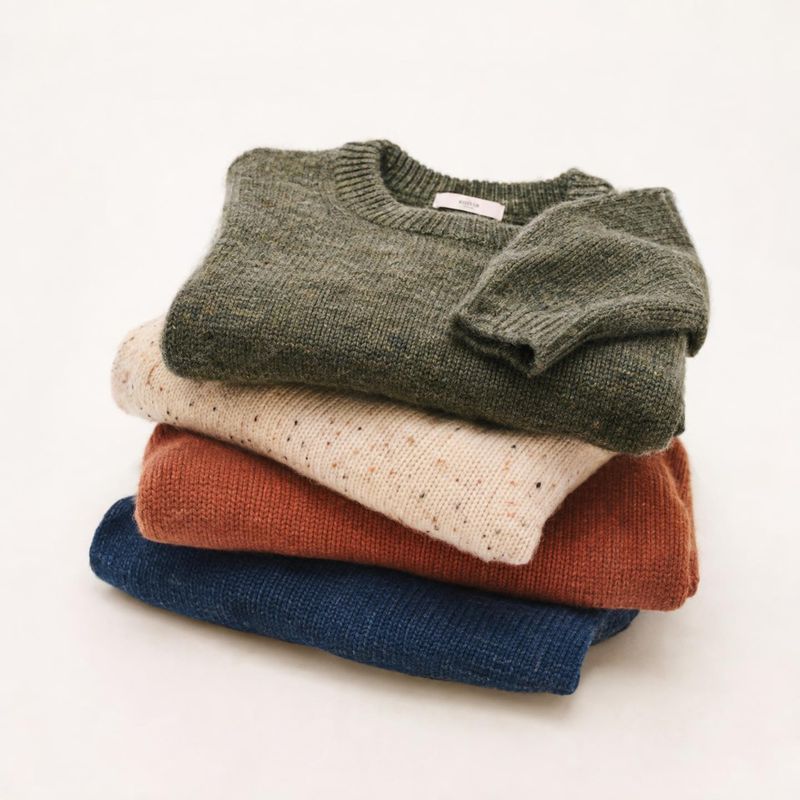 sweaters flat lay product photo: Four folded knit sweaters in muted green, cream, rust, and navy are stacked on a clean pale studio surface, with soft even lig…