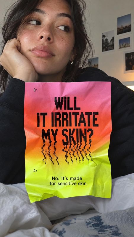skincare packet product in hand product photo: A woman in a black top holds a bright, crinkled skincare packet with sensitive-skin messaging close to the camer…