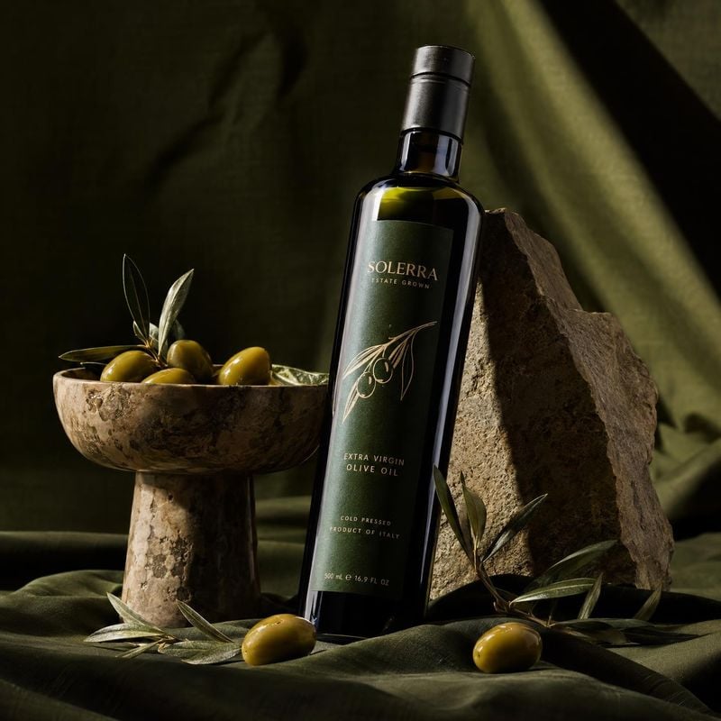 extra virgin olive oil black friday product photo: A dark glass bottle of olive oil stands among olives, olive branches, and rugged stone on deep green fabric,…