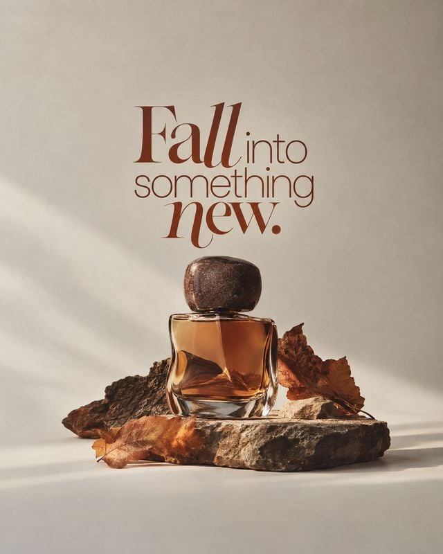 perfume bottle autumn product photo: An amber perfume bottle rests on weathered stone among russet leaves, lit by soft angled sunlight beneath elegant autumn-t…