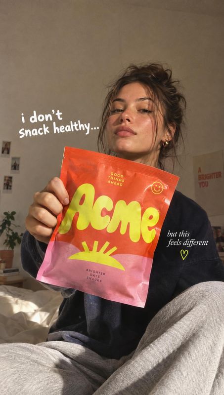 snack bag product in hand product photo: A woman in casual dark clothing holds a bright pink-and-orange snack bag toward the camera in a softly lit bedroom, wi…