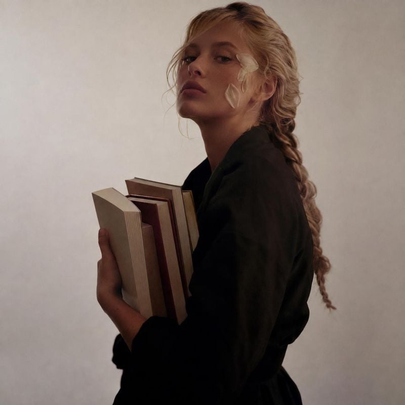 books book product photo: A blonde woman in a dark outfit holds several books against a softly lit neutral studio backdrop, creating a restrained, contemplativ…