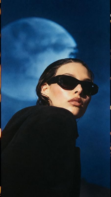 sunglasses on-body detail product photo: A model in black sunglasses and a dark outfit looks over her shoulder against a vivid blue sky, with dramatic low-angl…