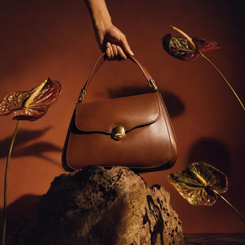 brown leather handbag product in hand product photo: A hand presents a structured brown leather handbag above a rugged rock, framed by glossy tropical leaves a…