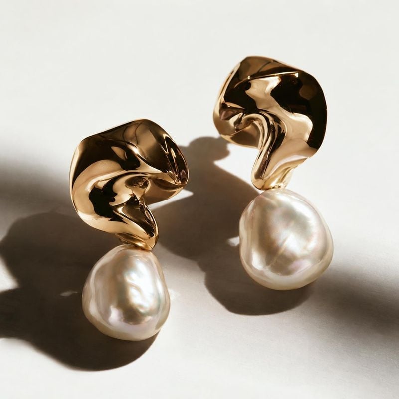 pearl drop earrings jewelry product photo: A pair of sculptural gold-tone earrings with luminous pearl drops rests on a pale surface, lit by warm directional l…