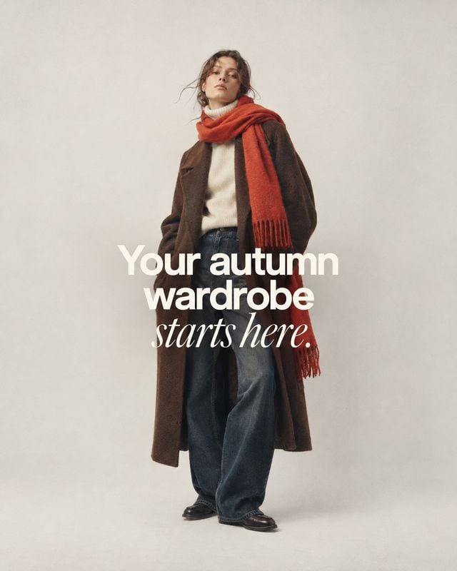autumn outfit autumn product photo: A model poses against a softly lit neutral studio backdrop in a long brown coat, cream sweater, dark jeans and a vivid rust…