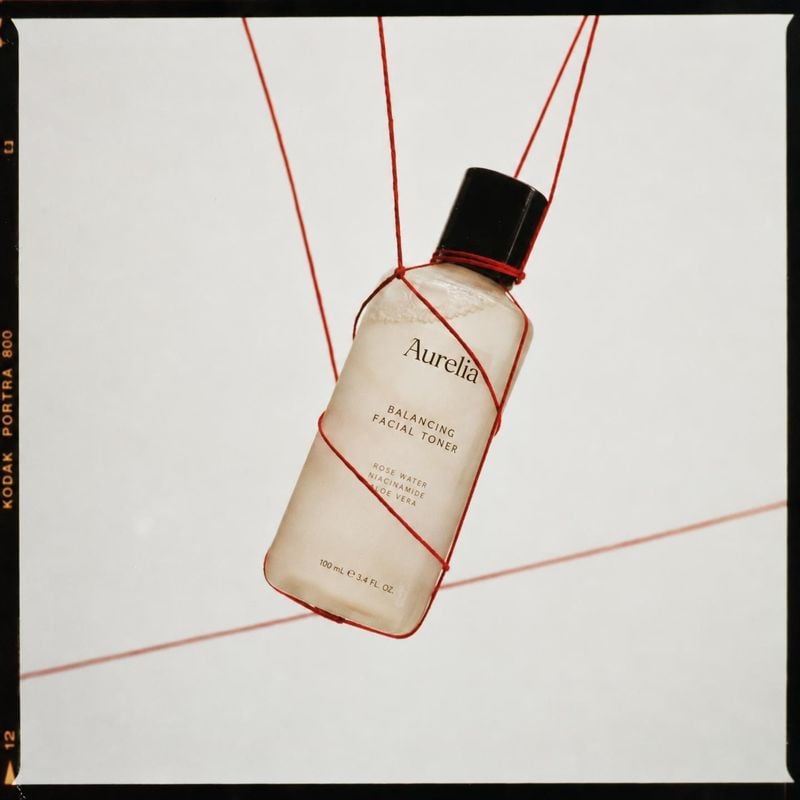 facial toner floating product product photo: A pale facial toner bottle with a black cap hangs at an angle from taut red strings against a clean white studio b…