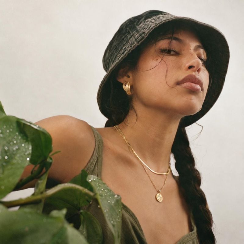 gold necklace and earrings on-body detail product photo: A model wears layered gold necklaces, hoop earrings, a dark bucket hat and an olive top against a soft…