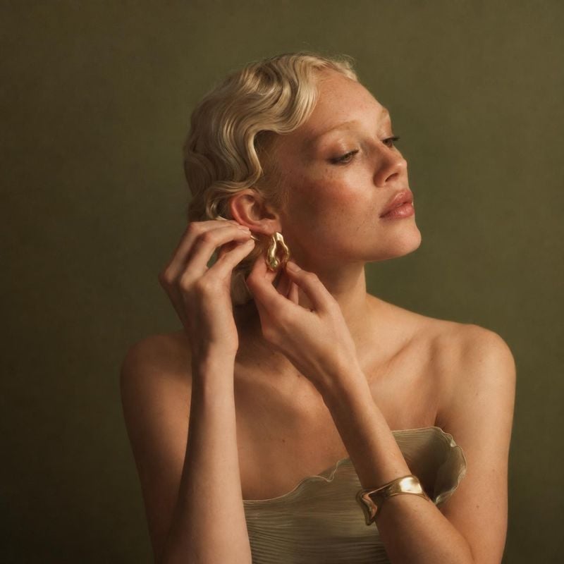 gold earrings and cuff bracelet on-body detail product photo: A short-haired model fastens a sculptural gold earring, wearing a matching cuff against a muted o…