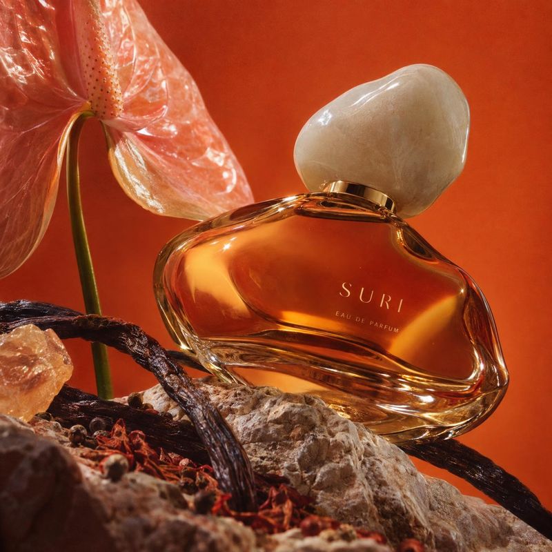 perfume ingredients product photo: An amber perfume bottle with a pale sculptural cap is arranged among flowers, dried botanicals and textured stones against a…