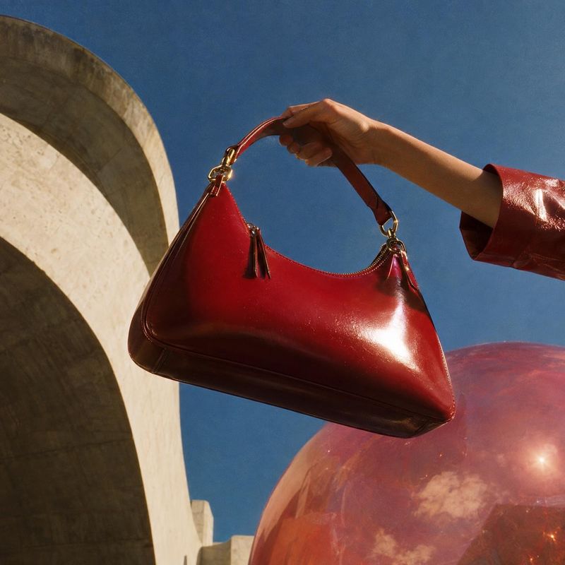 red leather handbag product in hand product photo: A glossy red handbag is lifted by its handle against a deep blue sky, with sculptural stone architecture and…