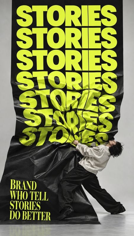 action & motion product photo: A person in a loose white top and dark trousers pulls at a crumpled black sheet covered in oversized neon-yellow lettering, phot…