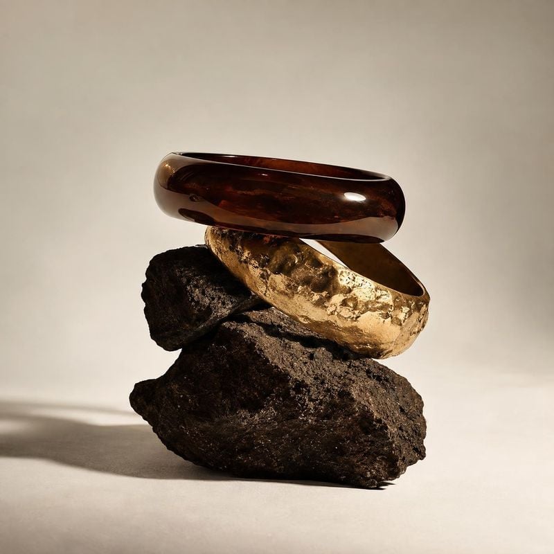 bracelets black friday product photo: Two sculptural bracelets, one glossy dark brown and one hammered gold, are stacked on rugged rocks against a warm neutral…