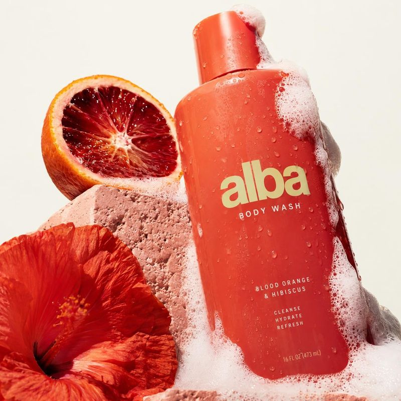 blood orange and hibiscus body wash beauty product photo: A coral body-wash bottle beaded with water and foam stands among a sliced blood orange and hibiscus f…