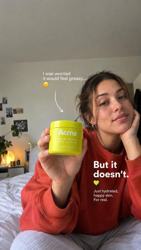 skincare cream lifestyle scene product photo: A woman in a red sweatshirt holds a bright lime-green skincare jar toward the camera in a softly lit bedroom, wit…