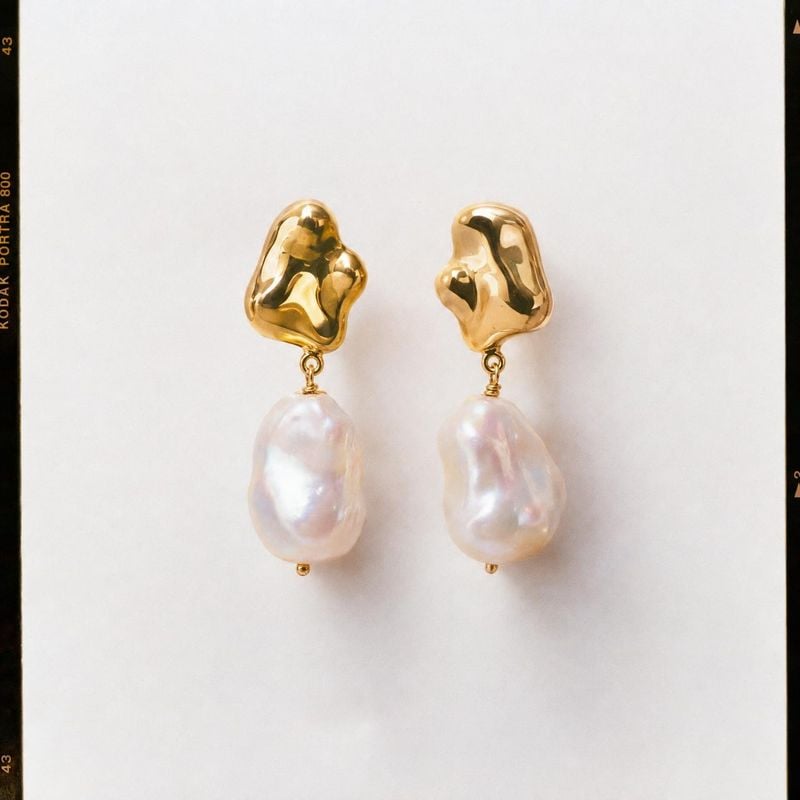 pearl drop earrings jewelry product photo: A pair of sculptural gold-tone earrings with luminous irregular pearls sits centered on a pale, softly lit backgroun…