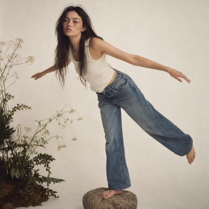 wide-leg jeans action & motion product photo: A model in a white tank and loose blue jeans balances barefoot on a rock against a warm neutral studio backdrop,…
