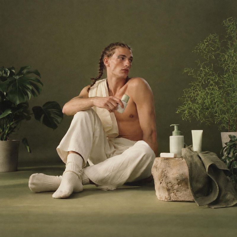body-care bottle lifestyle scene product photo: A man in relaxed cream clothing sits among leafy plants and understated toiletries, holding a small bottle in a…