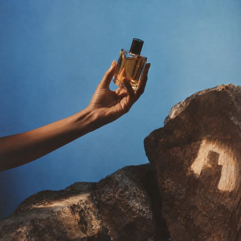 perfume product in hand product photo: A hand presents an amber perfume bottle against a vivid blue sky above rugged sunlit rocks, creating a warm, outdoorsy c…