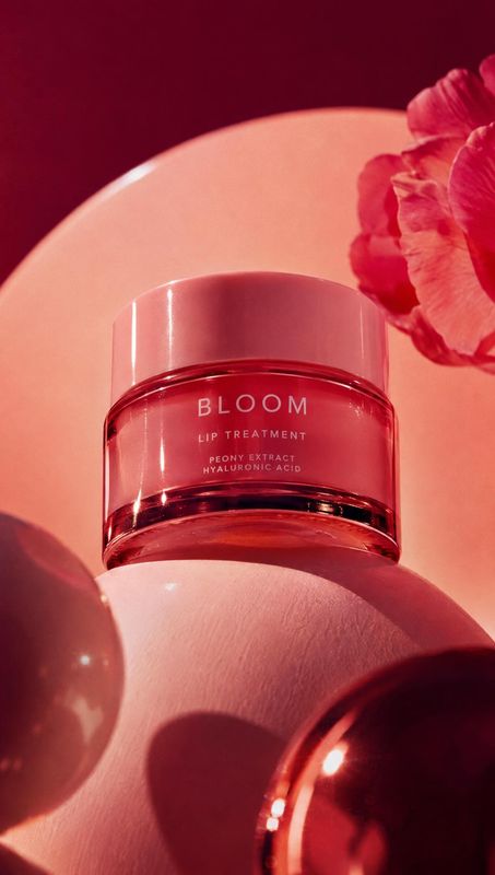 lip treatment close-up detail product photo: A red Bloom lip treatment jar rests on a sculptural pink surface beside a vivid flower, lit with warm saturated re…