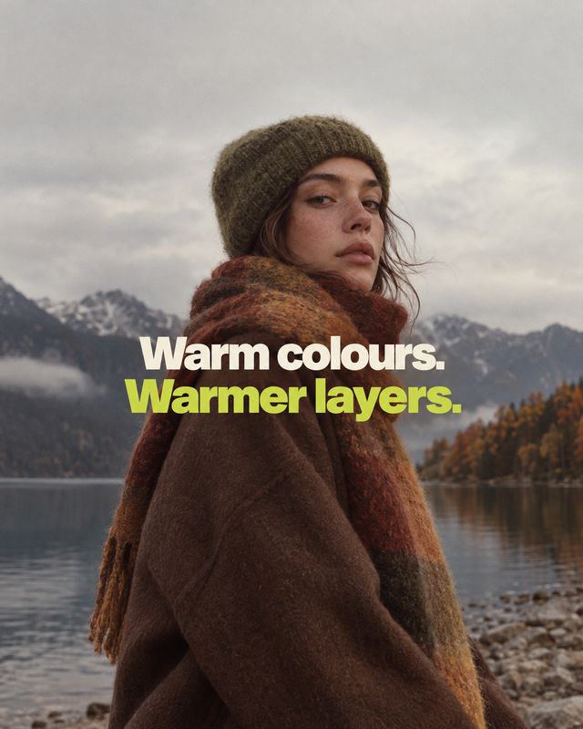 wool coat and scarf autumn product photo: A model in a brown wool coat, rust-toned scarf and knit beanie stands beside a mountain lake beneath cloudy autumn sk…