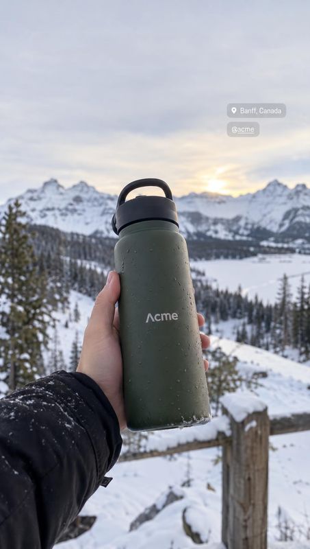 insulated water bottle product in hand product photo: A hand presents a dark green insulated bottle against a snowy mountain landscape at sunset, with a softly…