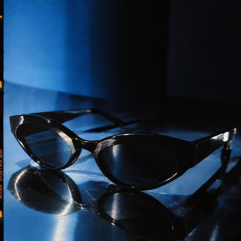 sunglasses close-up detail product photo: Black cat-eye sunglasses rest on a reflective blue surface, photographed in dramatic blue and black studio lighting f…