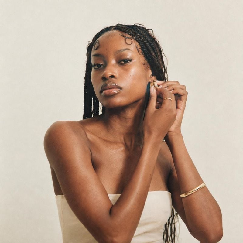 gold hoop earrings on-body detail product photo: A woman in a strapless cream top adjusts a gold earring against a softly lit neutral studio backdrop, creating…