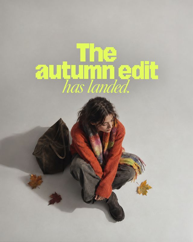 autumn outfit autumn product photo: A seated model in a rust-colored sweater, scarf, and relaxed trousers poses on a pale studio floor with a dark bag and scat…