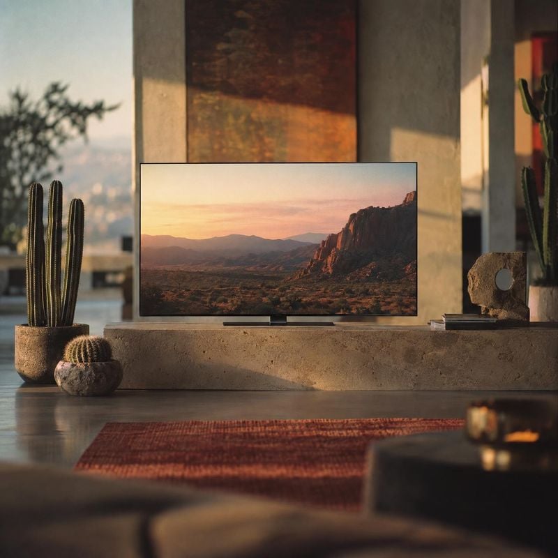 television lifestyle scene product photo: A slim television displaying a sunset-lit desert landscape sits on a stone console among cacti and decor in a warm, m…
