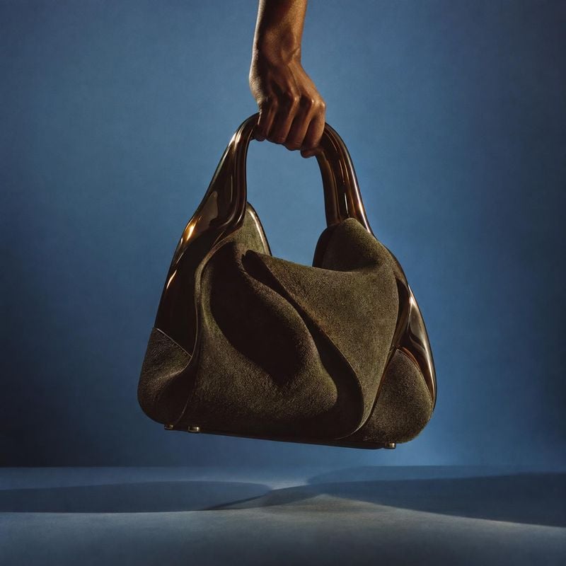 handbag product in hand product photo: A sculptural dark suede handbag with glossy curved handles is held aloft against a softly lit blue studio backdrop, crea…