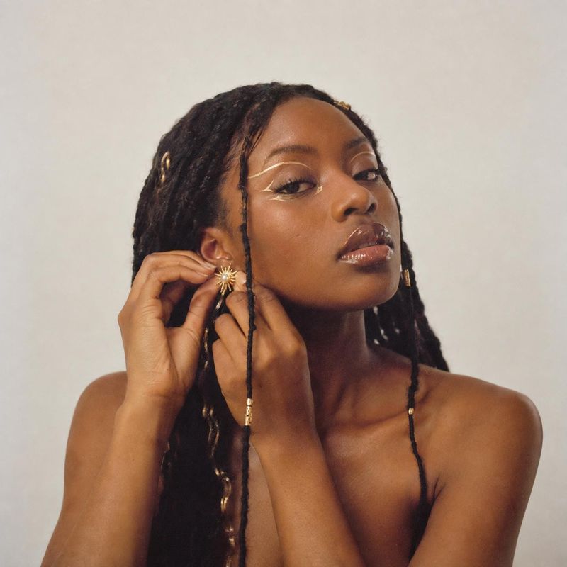 gold starburst earrings on-body detail product photo: A woman with long locs adjusts a gold starburst earring against a softly lit, neutral studio backdrop, cr…