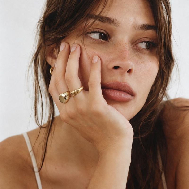 gold rings on-body detail product photo: A close beauty portrait shows a woman resting her hand against her freckled face, highlighting sculptural gold rings i…