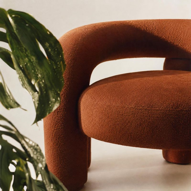 upholstered chair close-up detail product photo: A close view of a rust-colored upholstered chair beside glossy green leaves, lit with warm directional light a…